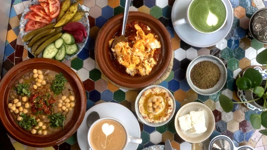 Frühstück von oben auf einem bunten Mosaiktisch: Shakshuka, Kichererbsen-Tajine, Hummus, Käse, Gemüse und Latte